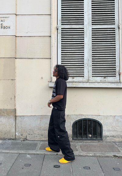 Persona con cabello rizado que lleva una camiseta gráfica negra, pantalones negros holgados y zapatos amarillos brillantes, caminando por la acera de la ciudad junto a una pared beige.