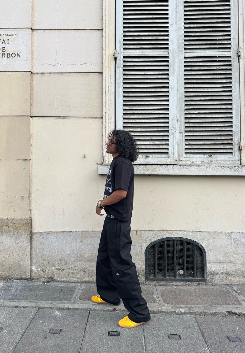 Personne avec des cheveux bouclés portant un t-shirt graphique noir, un pantalon noir ample et des chaussures jaunes vives marchant sur le trottoir d'une ville à côté d'un mur beige.