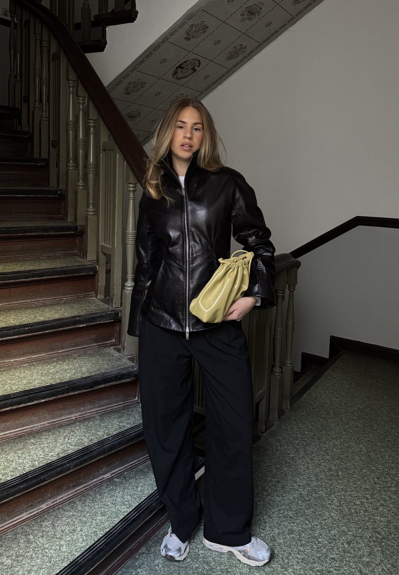 Jeune femme se tenant sur des escaliers, portant une veste en cuir noire, un pantalon noir large, des baskets blanches et tenant un sac à main beige froncé.