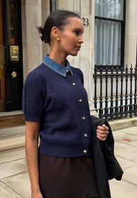 Granatowy kardigan z krótkim rękawem w splot ryżowy z kołnierzem z denimu, zdobiony srebrnymi guzikami. Nosi się go na brązową spódnicę, zabezpieczony ciemnym płaszczem.