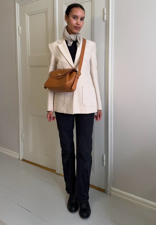Femme portant un blazer beige, une écharpe claire, un pantalon noir, des chaussures noires et un sac bandoulière en cuir marron, debout à l'intérieur près d'une porte blanche.