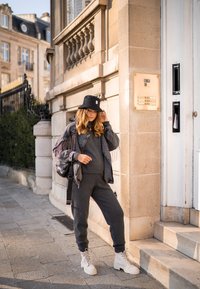 Gray knit jumpsuit with a distressed black jacket featuring fringe details. Black bucket hat and white lace-up boots complete the outfit.
