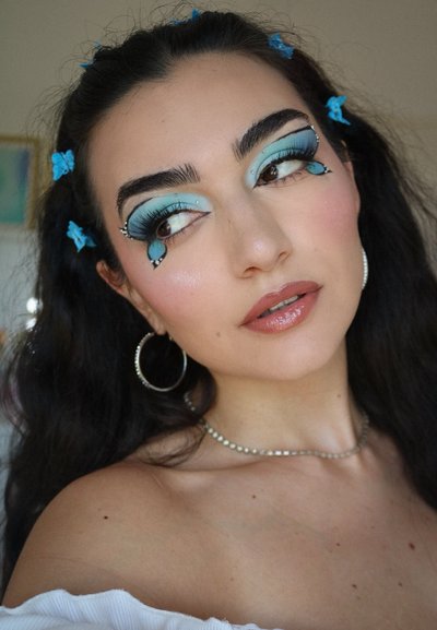 Maquillaje que presenta sombras de ojos azules con diseños de lágrima, complementado con aretes de aro y una cadena de perlas, todo sobre un fondo desenfocado.