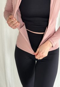Világos rózsaszín cipzáras kabát hosszú ujjal, két elülső zsebbel, fekete ujjatlan top és fekete leggings fölött viselve. Az anyag simának és testhezállónak tűnik.
