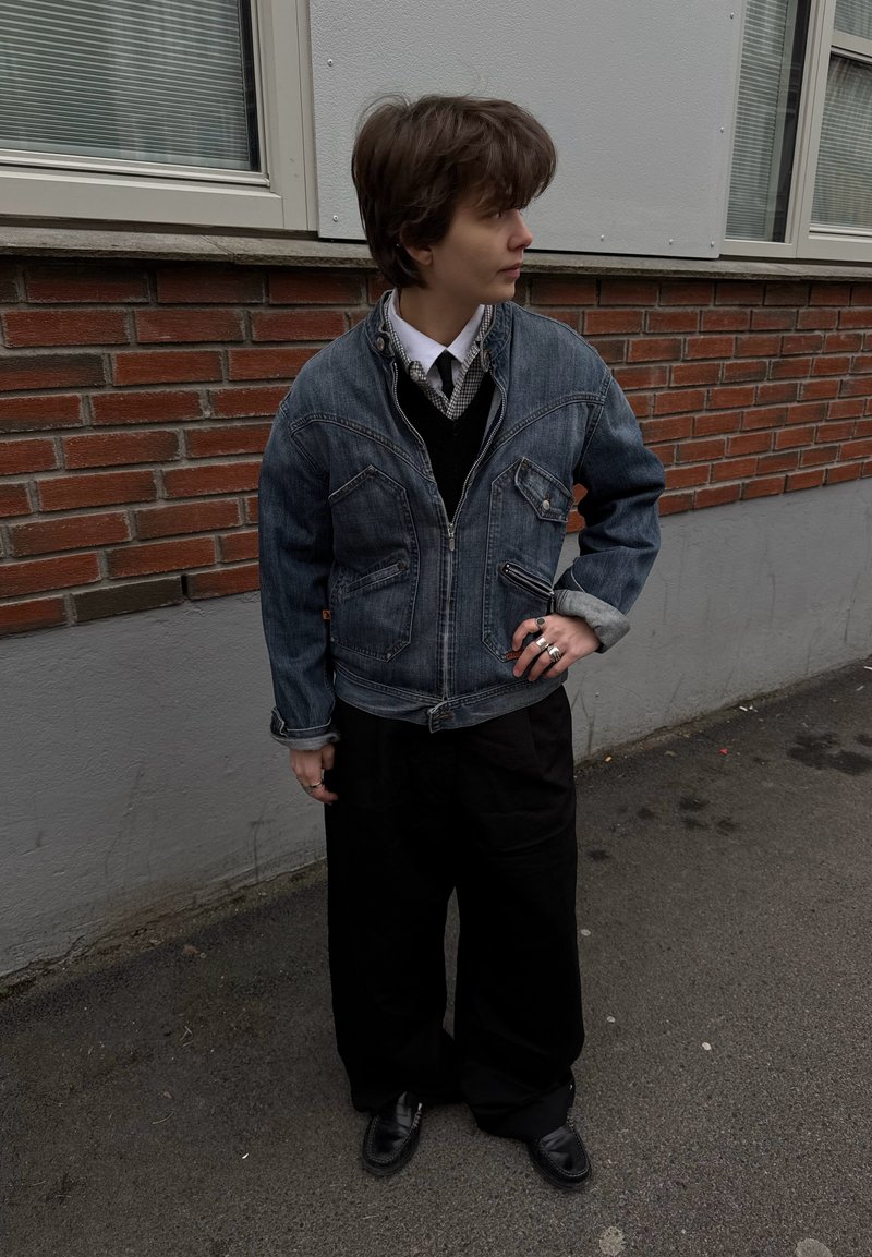 Jeune personne debout sur le trottoir portant une veste en jean, un pantalon large noir, des chaussures noires et une chemise blanche avec cravate, à côté d'un bâtiment en briques.