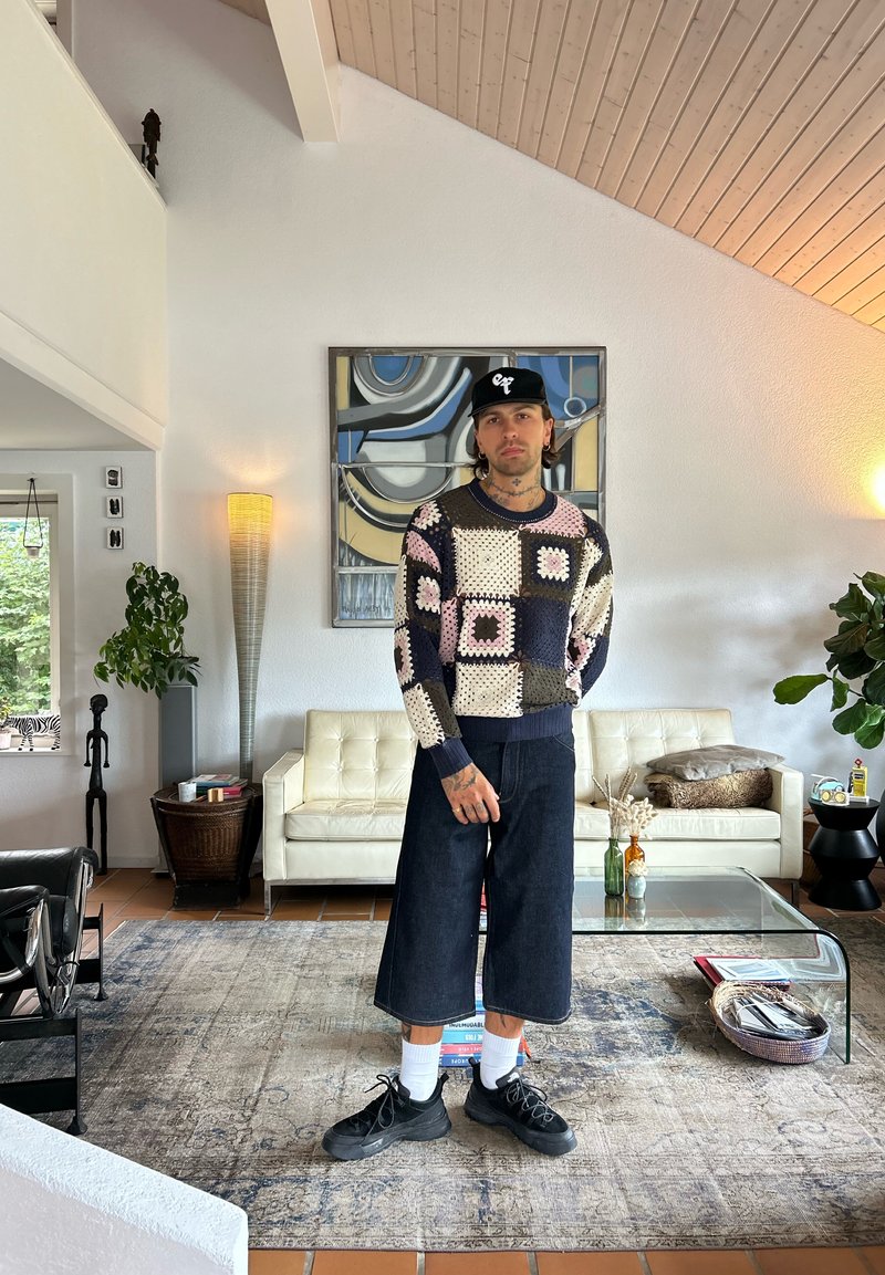 Uomo che indossa un maglione a fantasia, culotte in denim, sneakers nere e un cappellino nero, si trova in un soggiorno moderno con un tavolino da caffè in vetro.