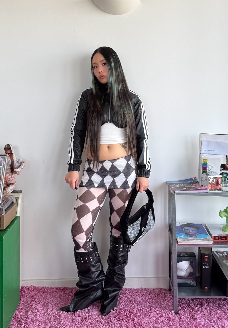 Jeune femme aux longs cheveux foncés debout à l'intérieur, portant une veste noire courte, une jupe à motif losange et des leggings, des bottes noires souples, tenant un sac.