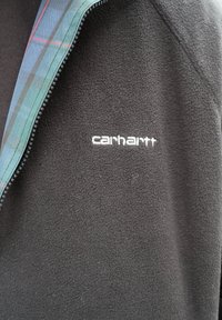 Veste polaire noire avec une fermeture éclair à carreaux bleu et vert partiellement ouverte et le logo "Carhartt" brodé en blanc sur la poitrine.