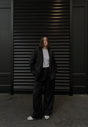 Blazer negro oversize, pantalones de pierna ancha a juego y camiseta blanca. La persona está de pie contra un fondo negro texturizado, vistiendo zapatillas en gris y blanco.