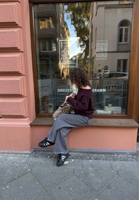 Pull marron oversize, jean gris à jambes larges, baskets noires avec des rayures blanches, tenant un sac à motif, assis sur un rebord en bois.