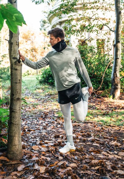 Una persona se estira contra un árbol, vistiendo una camiseta de manga larga gris claro, pantalones cortos negros y leggings blancos con acentos amarillos.