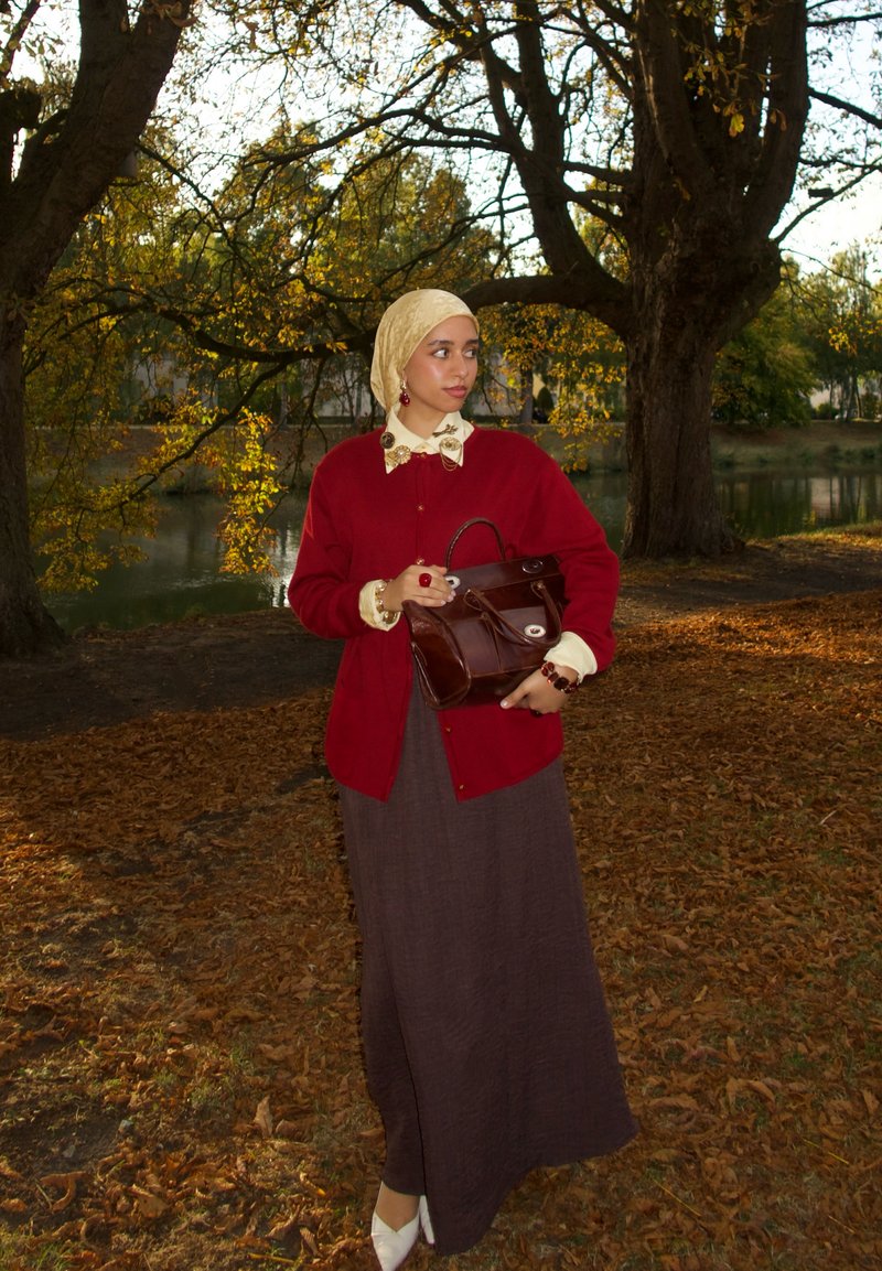 Cardigan rosso sopra una blusa crema con fiocco, gonna lunga marrone, scarpe bianche e borsa marrone. Foulard in testa con motivo dorato e gioielli visibili.