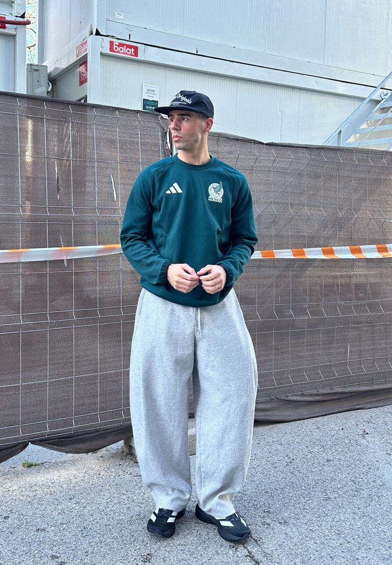 Homme portant un sweat-shirt vert du Mexique, un pantalon large gris, des baskets noires et une casquette noire, debout devant une barrière de chantier sur le trottoir.