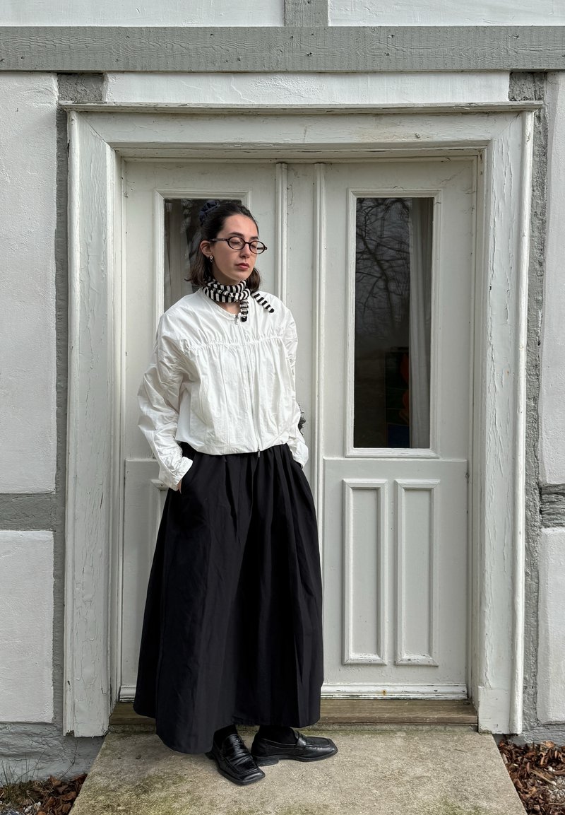 Casaco branco, leve, com fecho éclair e detalhes franzidos, combinado com uma saia longa preta. Usado com um lenço listrado em preto e branco e sapatos.