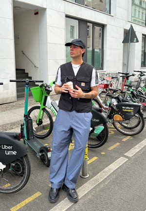 Chaleco negro con cremallera sobre una camiseta blanca, pantalones anchos azules y zapatos negros. Sosteniendo una taza de café con bicicletas y patinetes de fondo.