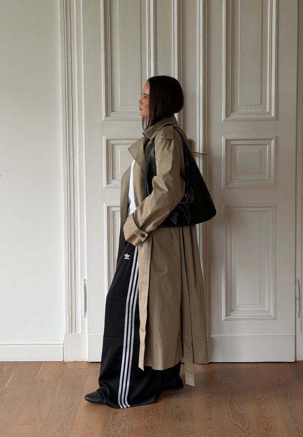 Mujer con abrigo beige, pantalones deportivos negros Adidas y bolso negro al hombro, de pie de lado frente a puertas blancas con paneles sobre suelo de madera.