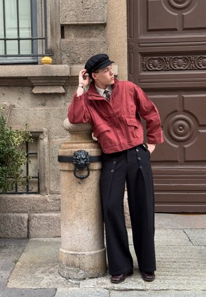 Red cropped jacket with two front pockets, black wide trousers with large buttons, and black cap; standing next to a stone pillar.