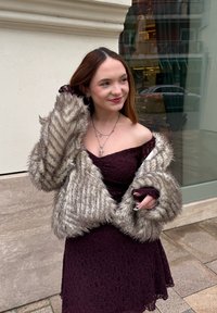 Fiatal nő bordó váll nélküli csipke ruhában és szürke műszőrme kabátban áll a szabadban, egy üveges ablakú épület közelében.