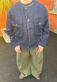 Farmerdzseki két elülső zsebbel, ezüst patentgombokkal és kontrasztos varrással, laza szabású olívazöld nadrággal párosítva. Természetes anyagtextúra.