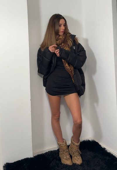 Chaqueta negra acolchada sobre un vestido mini negro ajustado, combinada con botas marrones forradas de piel y una bufanda marrón con estampado. Sobre un fondo liso.