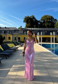 Abito in satin rosa con scollatura a cappuccio e spalline sottili. Sullo sfondo si vede una piscina, un patio e un edificio giallo con alberi.