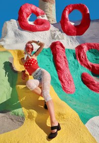 Rdeče kopalke z belimi pikami, bele čipkaste pajkice in črni čevlji z visokimi petami. Barvna teksturirana stena z rdečo, belo, zeleno in rumeno barvo.