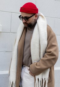 Béžový vlnený kabát cez hnedý rolák, biely šál s strapcami, červená pletená čiapka, čierne slnečné okuliare a biele nohavice.