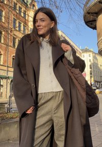 Une femme porte un long manteau marron foncé à larges manches par-dessus un pull gris clair et un pantalon rayé gris, portant un sac matelassé marron.