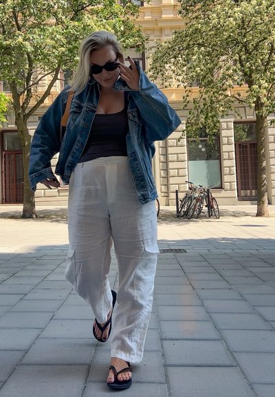 Chaqueta de denim, top negro de tirantes, pantalones cargo de lino blancos y chanclas negras. Entorno urbano con árboles y bicicletas en el fondo.