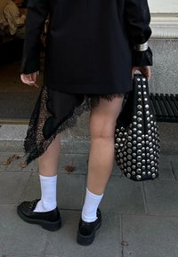 Czarna marynarka nałożona na spódnicę z koronkowym wykończeniem, w połączeniu ze zdobioną czarną torbą i czarnymi butami. Widoczne białe skarpetki do kolan.