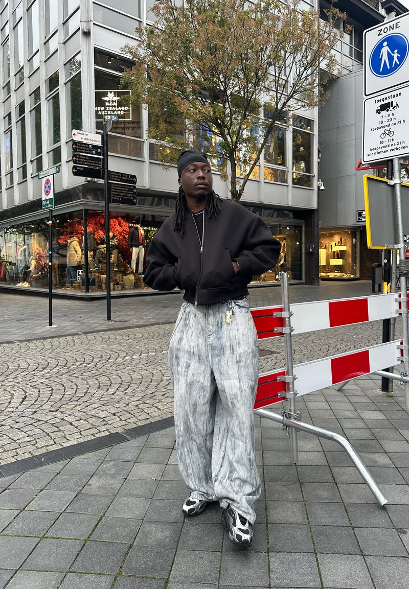 Svart zip-up bomberjacka, grå mönstrade over-size byxor och svarta sneakers med unik design, står nära en byggbarriär.