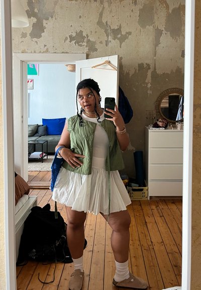 Chaleco acolchado verde sobre un vestido blanco con falda acampanada. Cabello trenzado, gafas y zuecos beige. Suelo de madera clara en una habitación.