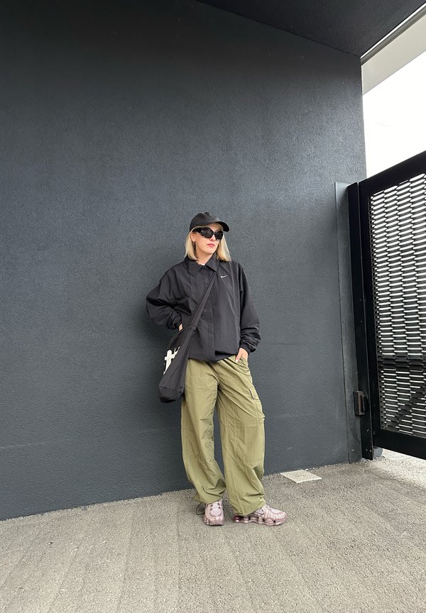Casaco oversized preto, calças cargo verdes-oliva, ténis cor-de-rosa e um boné preto. Os acessórios incluem óculos de sol pretos e uma pequena bolsa preta.
