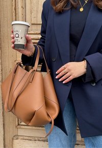 Femme tenant un sac à main en cuir marron et un gobelet de café en papier, portant un blazer bleu marine, un haut noir, des bijoux en or et un jean bleu.