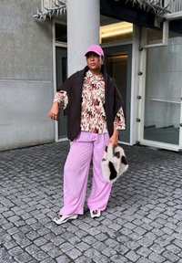 Volné růžové pruhované kalhoty, krémová košile se zvířecím vzorem, hnědé oversized svrchní oblečení, růžová čepice a kabelka s chlupatým vzorem. Pozadí z kamenů.