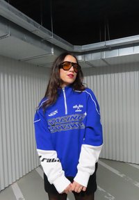 Modrý a bílý pulovr na zip s nápisem "RACING" ve zlaté barvě. Má dlouhé rukávy a vysoký límec, nošený přes černou sukni.