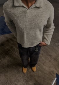 Personne portant un pull texturé crème, un jean foncé avec un porte-clés, et des chaussures marron clair, debout sur un sol moucheté avec des marques bleues.