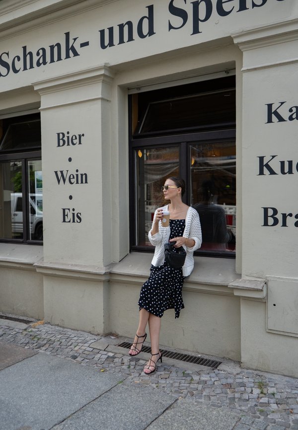 Modelo com um vestido preto de bolinhas e uma fenda, cardigan de malha branco e sandálias com tiras, segurando uma bebida, sentada à janela de um restaurante.