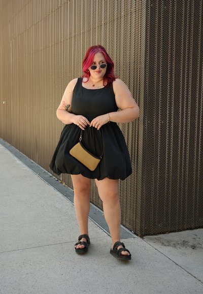 Vestido negro sin mangas con falda acampanada, acompañado de un bolso de mano dorado y sandalias negras. Cabello largo y ondulado con mechas rosas.