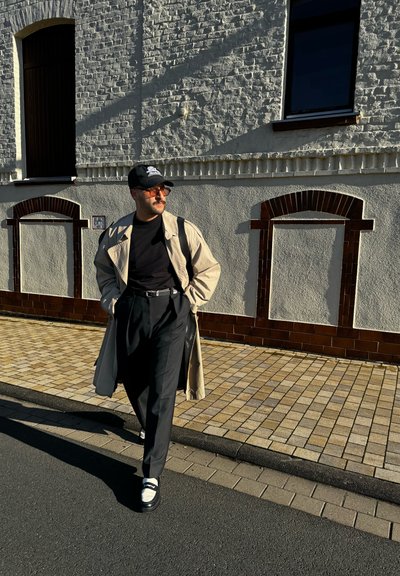 Gabardina beige sobre una camiseta negra, pantalones negros y zapatos en blanco y negro. Gafas de sol y una gorra negra. Calle pavimentada y fondo de edificios.