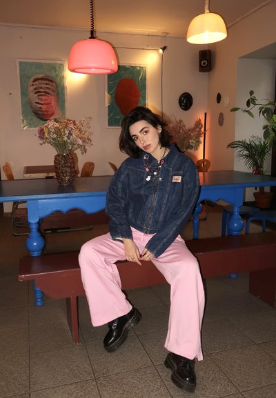 Chaqueta de mezclilla con cremallera delantera, pantalones de pierna ancha rosas y botas negras. Arte abstracto en la pared y una lámpara colgante rosa en un ambiente de café.