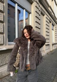 Veste en fausse fourrure de couleur gris foncé, avec un large col, une coupe ample et un tissu texturé. Associée à un jean gris et un sac à motifs.