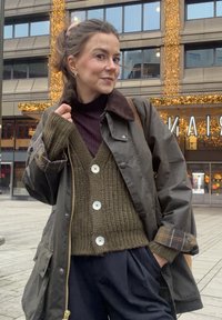 Olijfgroene, wollen gilet met witte knopen, gelaagd over een donkere coltrui, gecombineerd met een donkergroene, waterdichte jas en marineblauwe broek.