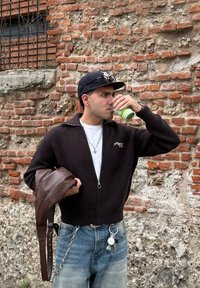 Sötétbarna bordázott zipzáras dzseki, alatta fehér póló, világoskék farmer, zöld ital a kezében, barna bőrtáska és kulcstartó.