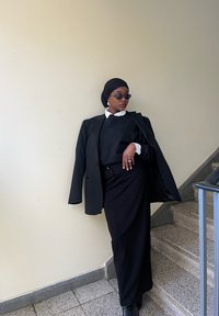 Schwarzer maßgeschneiderter Blazer über einem schwarzen Pullover und einem weißen Hemd mit Kragen, kombiniert mit einem schwarzen Rock, akzentuiert durch Sonnenbrillen und ein Kopftuch.