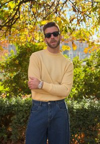 Άνδρας που φοράει κίτρινο πουλόβερ, σκούρα γυαλιά ηλίου και μπλε τζιν στέκεται έξω με σταυρωμένα τα χέρια κάτω από ένα φθινοπωρινό δέντρο με κίτρινα φύλλα.