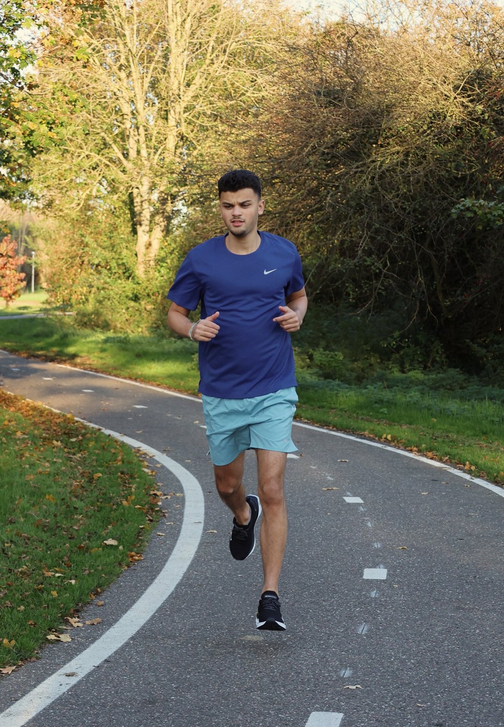 Maglietta atletica blu e pantaloni corti azzurri, che corrono su un percorso curvo circondato da vegetazione. Scarpe da corsa nere; design casual e sportivo.