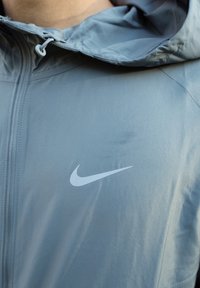 Casaco com capuz da Nike em tecido cinza claro, apresentando um fecho frontal e um logotipo swoosh branco no peito. Textura suave, design casual.