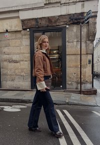 Bruine cropped jas met een witte kraag, donkere wijde jeans en bruine haklaarzen. Houdt een bruine tas vast en staat naast een stenen muur.