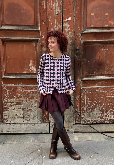 Mujer con cabello rizado y rojo, vestida con suéter estampado, falda plisada, medias y botas, de pie frente a grandes puertas de madera envejecida.